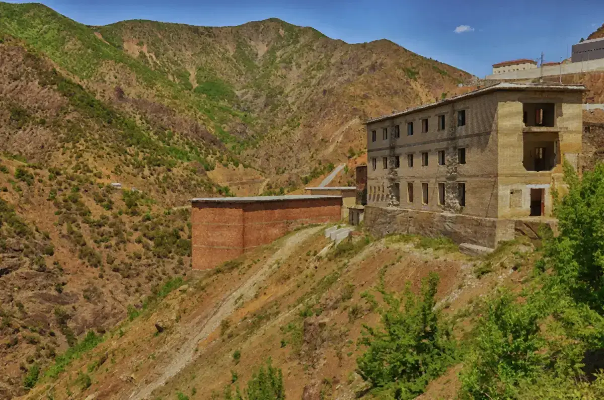 Ruined concrete and brick prison buildings on a barren, terraced mountainside under a blue sky