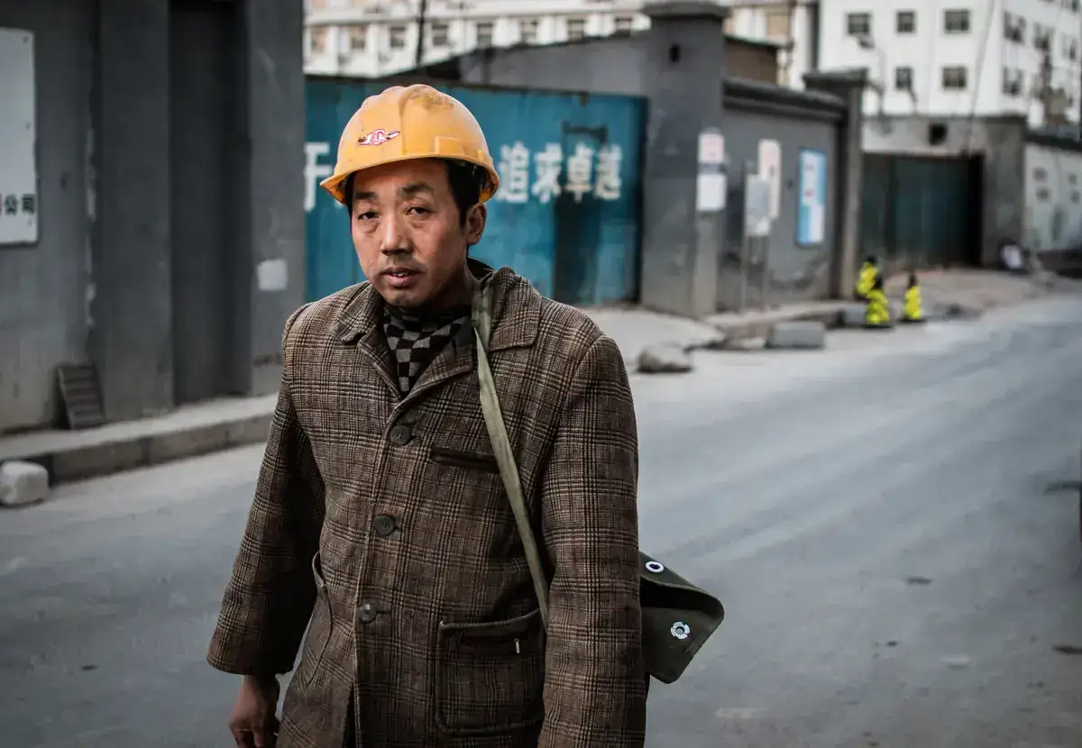 A middle-aged man in a yellow hard hat and a worn tweed jacket, a canvas bag over his shoulder, walking along a street past construction hoardings