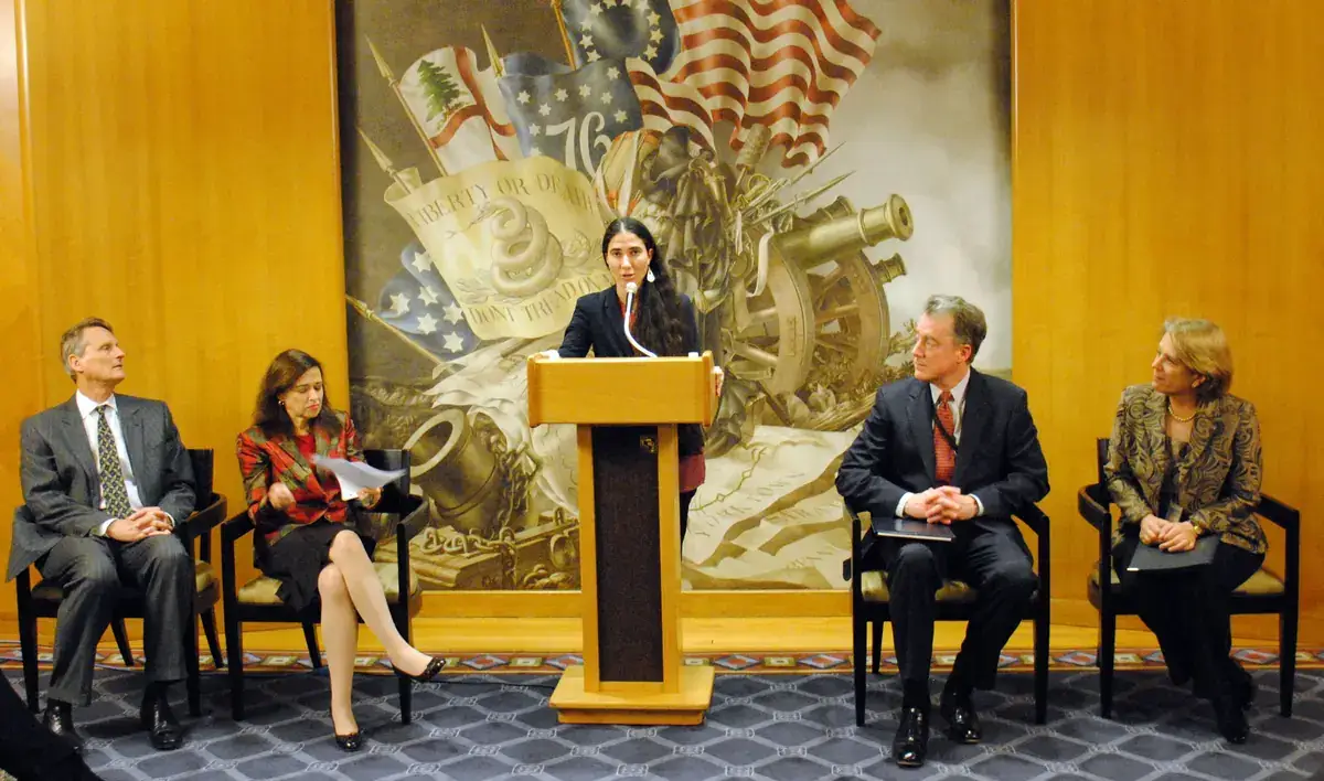 Yoani Sánchez speaking at a lectern with microphones, in front of the flags at the U.S. Department of State