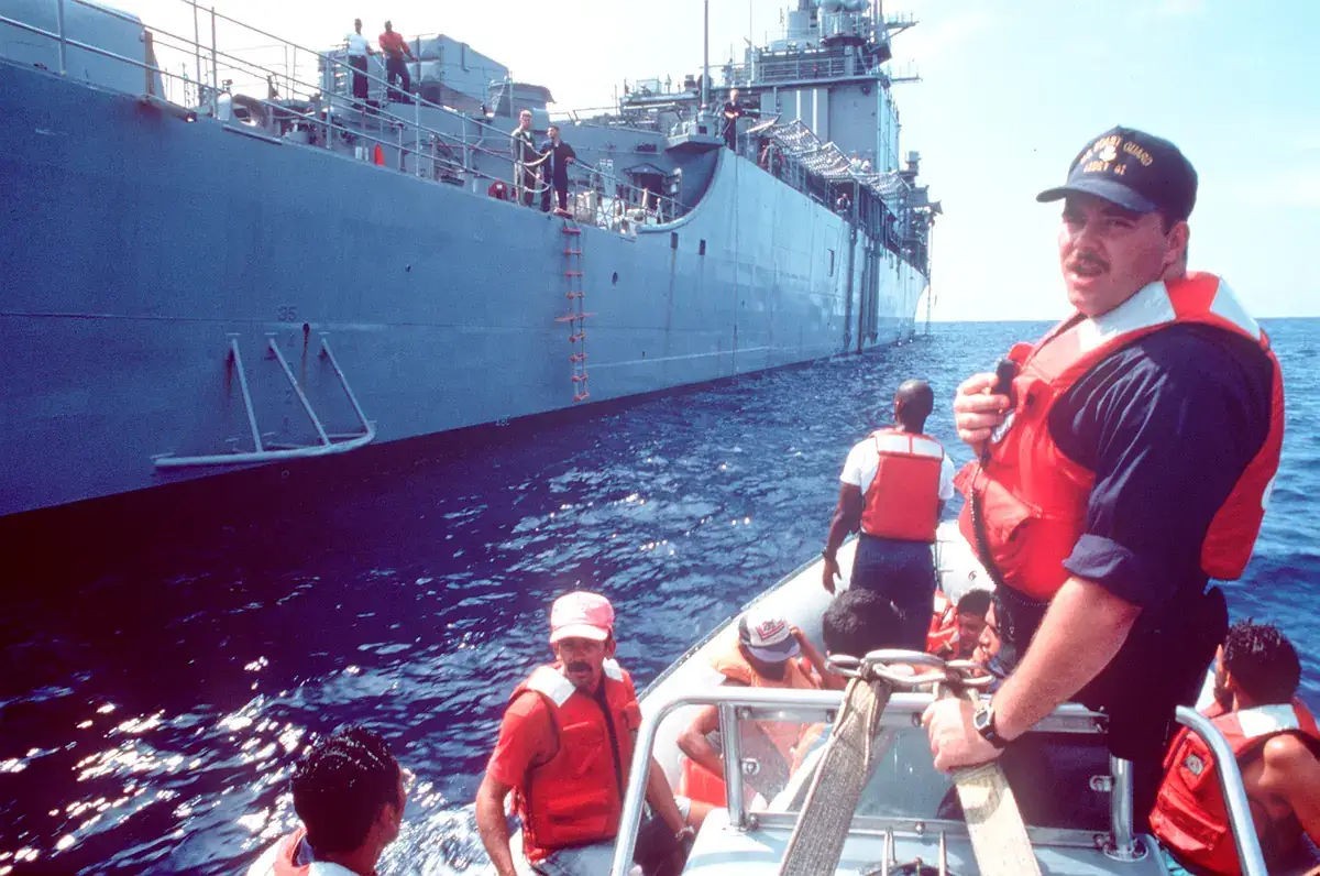 A U.S. Coast Guardsman in a life jacket stands in a small boat carrying Cuban migrants in orange life jackets alongside a grey U.S. Navy ship at sea