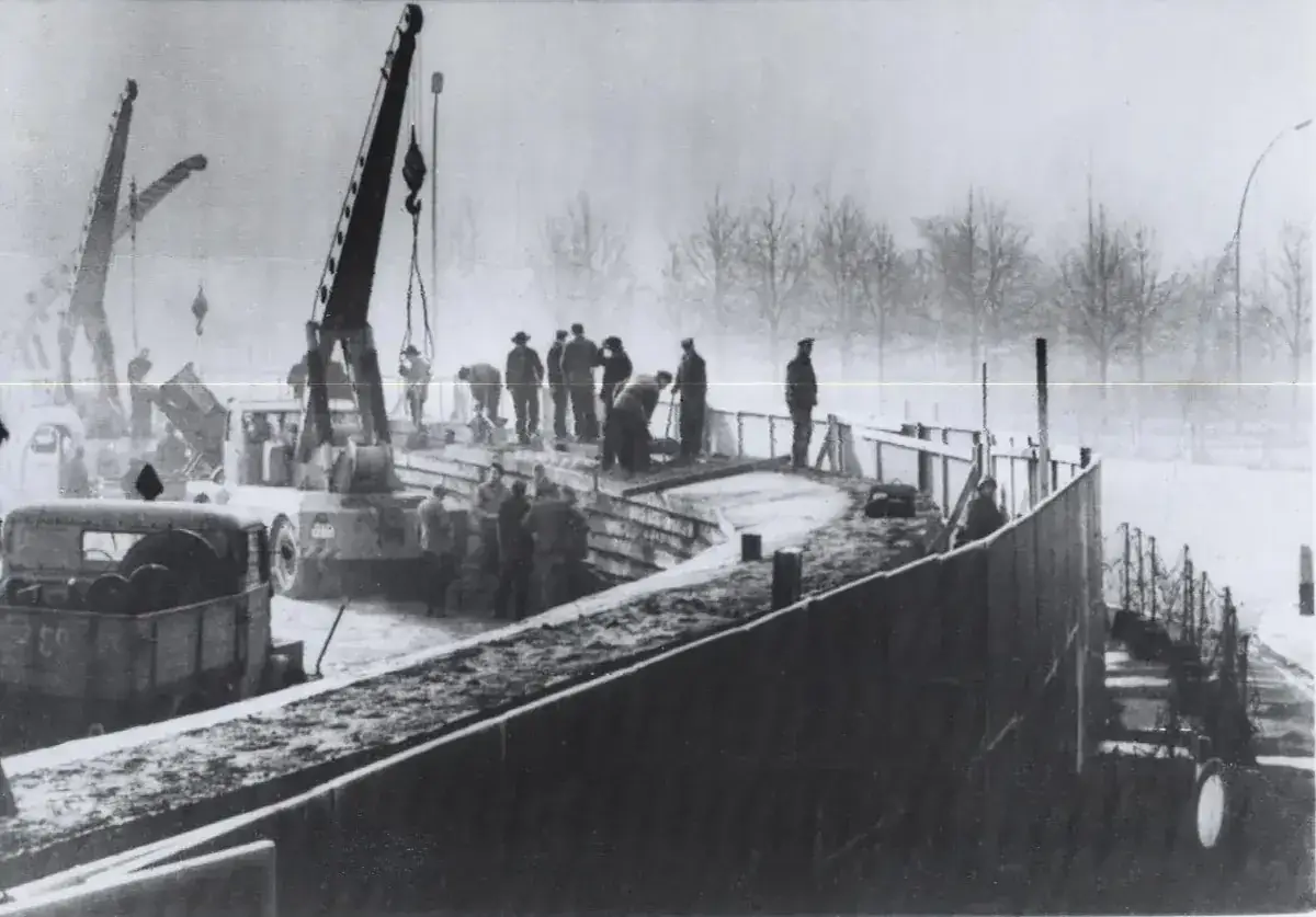 Black-and-white photograph of workers and cranes building a concrete wall on a snowy street, with bare trees in the fog behind