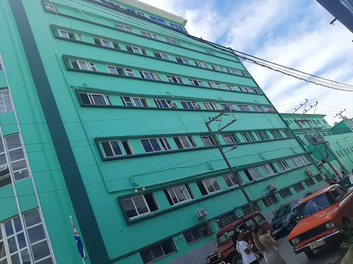 A large turquoise-painted hospital building with rows of windows, seen from the street with old cars parked in front and a Cuban flag at the entrance