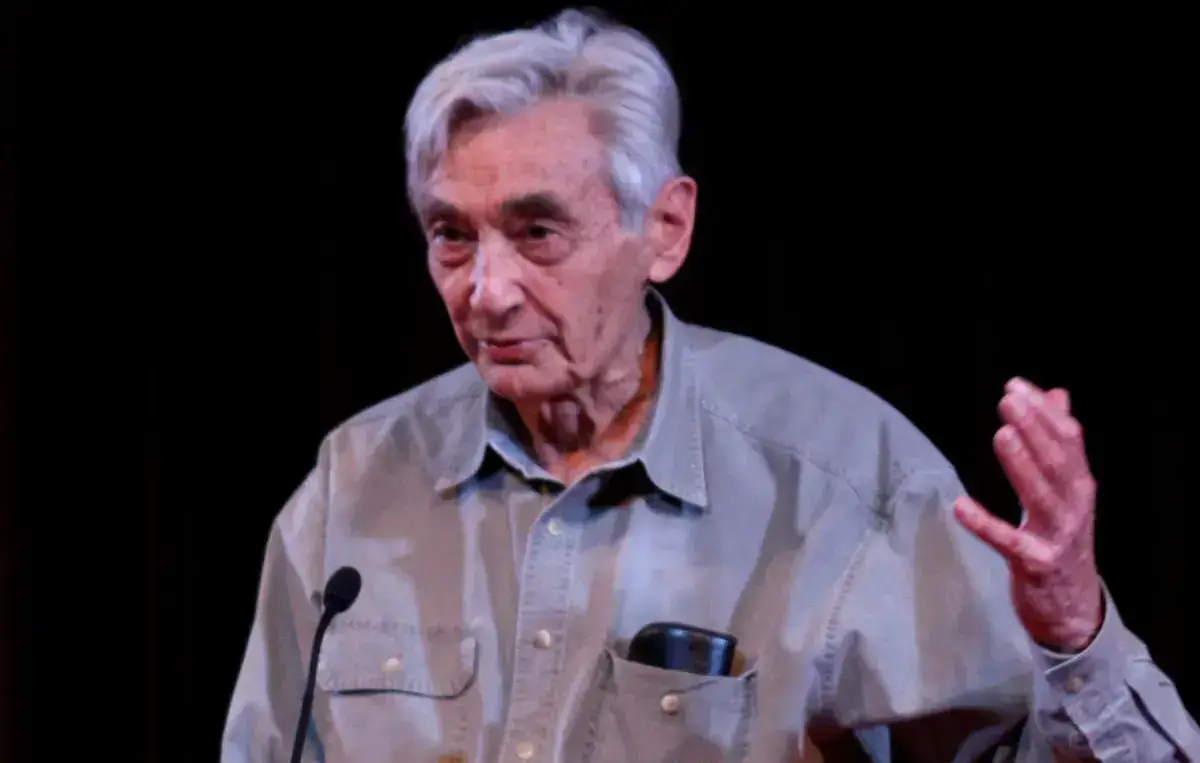 Howard Zinn, white-haired and in a grey shirt, gesturing with one hand as he speaks at a microphone against a dark background