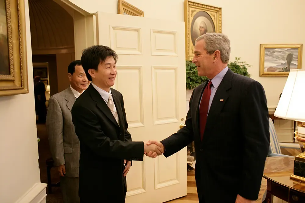 President George W. Bush shaking hands with Kang Chol-hwan in the Oval Office, with an older man standing behind Kang