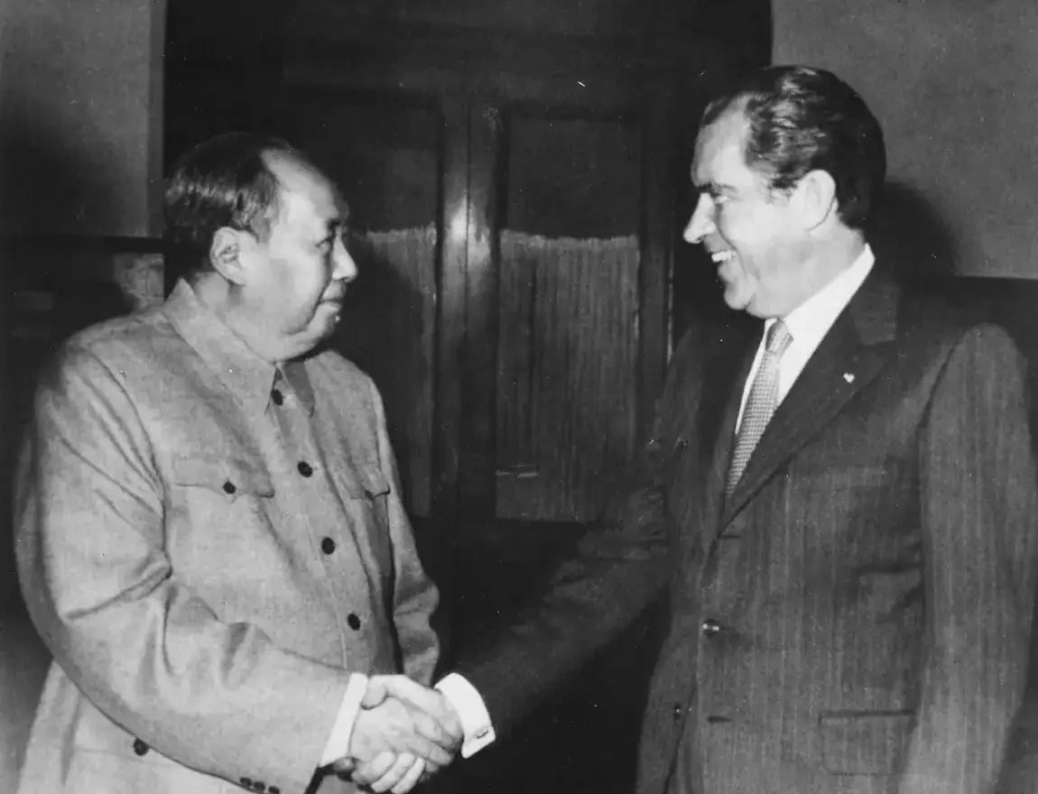 Black-and-white photograph of Mao Zedong, in a grey buttoned tunic, shaking hands with a smiling Richard Nixon in a dark suit, in a wood-panelled room