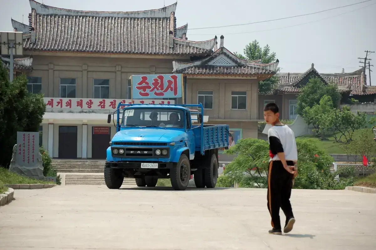 A blue truck parked before a traditional-style building with red Korean signs, as a boy walks across the forecourt, at a farm in North Korea