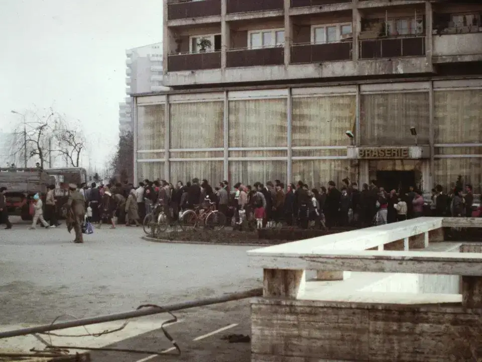 A long queue of people in winter coats, some with bicycles and shopping bags, along the front of a concrete apartment block in Bucharest
