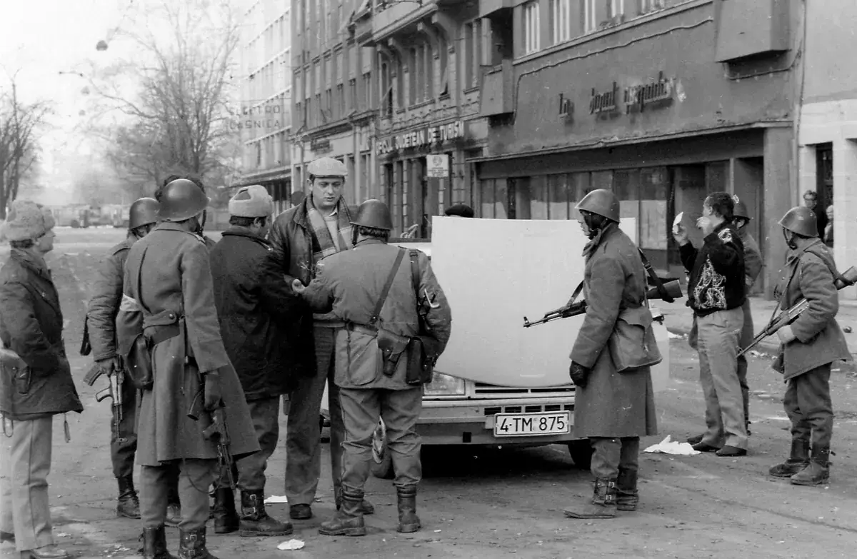Black-and-white photograph of soldiers in helmets with rifles and civilians gathered around a car with its bonnet raised on a city street in Timișoara
