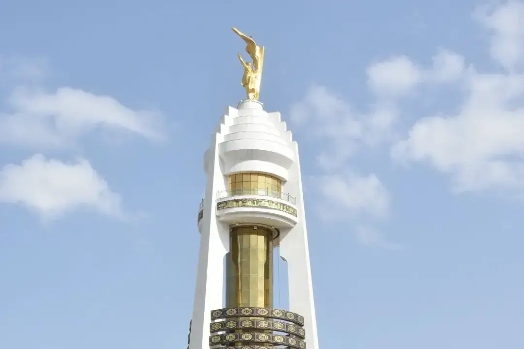 The top of a tall white tower with gold-banded sections, crowned by a gilded statue of a figure with raised arms, against a blue sky
