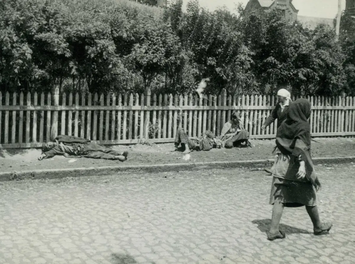 Three emaciated people lying and sitting on the pavement beside a wooden fence on a Kharkiv street, as two women walk past