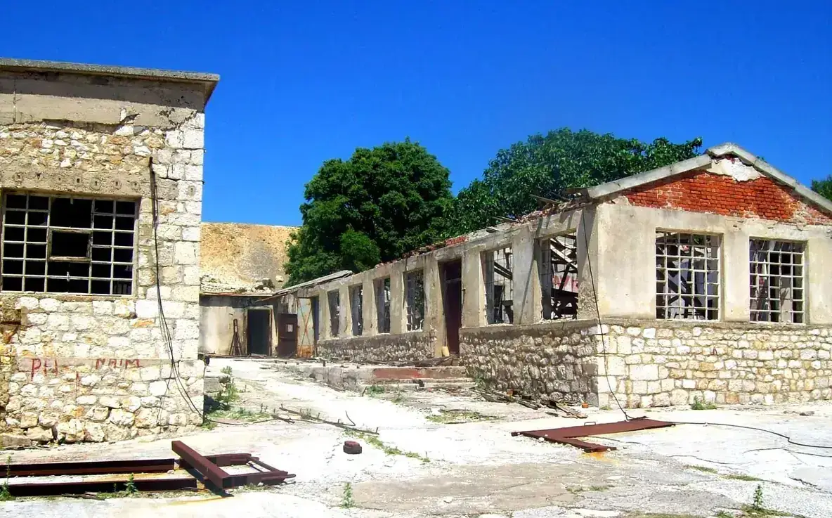 Abandoned stone prison barracks with barred windows and a collapsing roof on the island of Goli Otok, under a blue sky